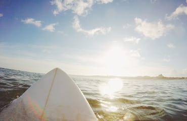The tip of a surfboard protrudes out over open ocean with the sun shining down.