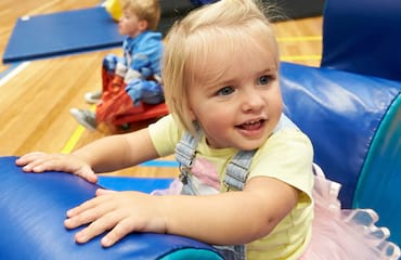 A blonde little girl smiles as she climbs over a soft cylinder climbing toy.