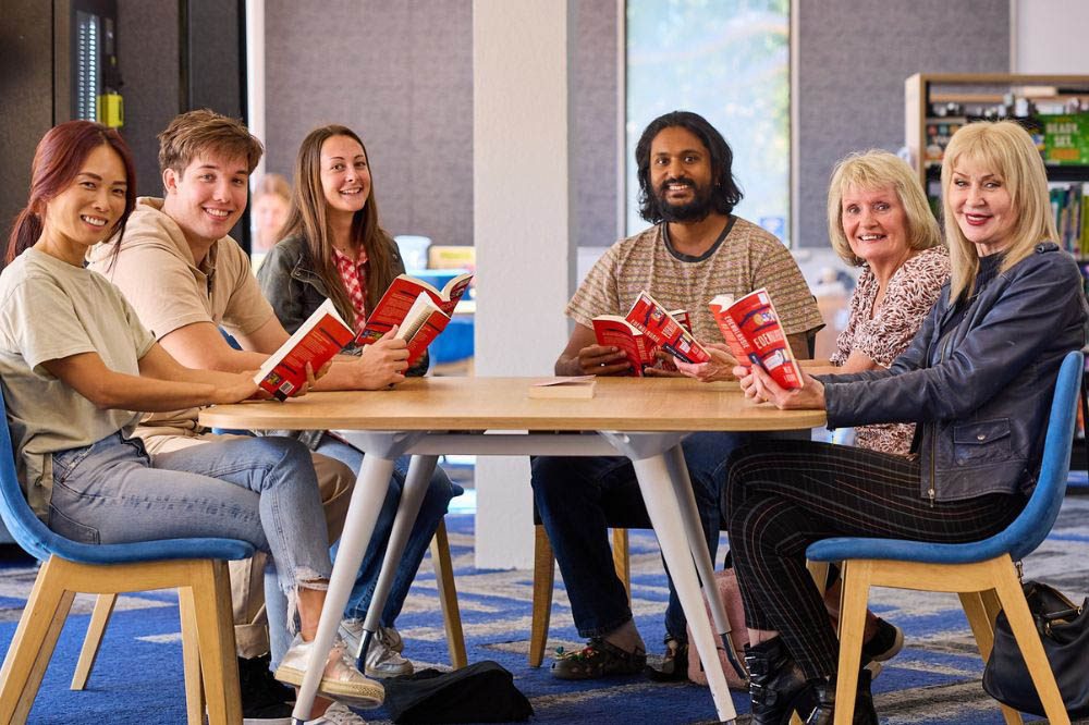Group of people sitting around a table, smiling and reading books together. 