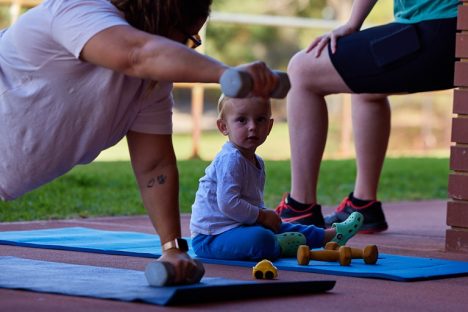 Two people exercising outside on mats and a baby watching one of them.