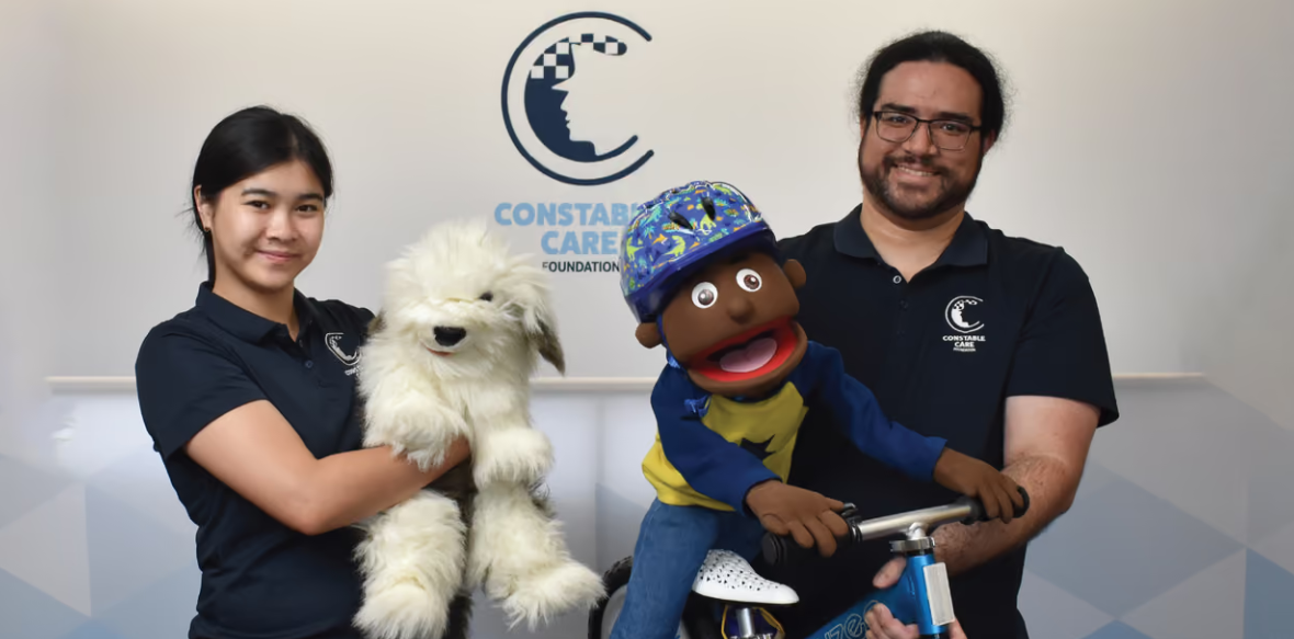 Two people smiling holding puppets, one is a dog puppet the other a puppet on a bike.